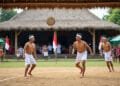 Ilustrasi tiga pemuda mengenakan pakaian tradisional dan kepala ikat sedang bertanding Sepak Takraw di lapangan beralaskan tanah dengan latar bangunan beratap rumbia serta bendera Indonesia